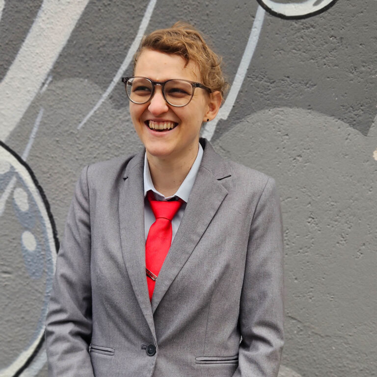 KLara Schinnerl, dunkeblonde kurze lockige Haare, Brille, steht in grauem Anzug mit roter Krawatte fröhlich lachend vor einer mit grauen Farben besprühten Wand